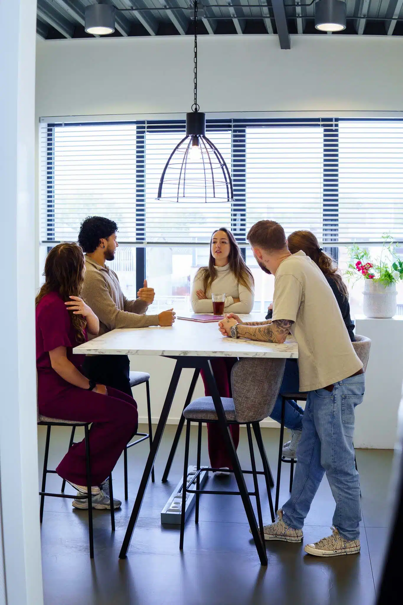 BURO210 Fullservice Marketingbureau Team van diverse collega's in moderne kantoorruimte met grote ramen, vergaderend rond een vierkante tafel met een marmeren blad en bespreken ideeën in een informele setting.