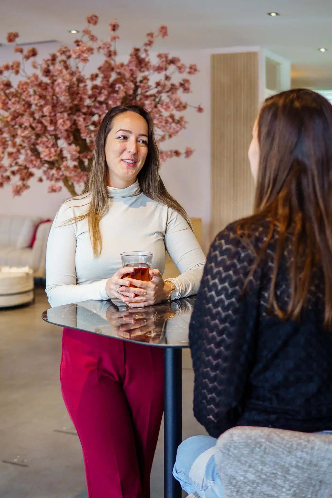 BURO210 Fullservice Marketingbureau Twee vrouwen in gesprek in een stijlvolle lobby, één met een glas thee, rode broek, en een lachende uitdrukking.