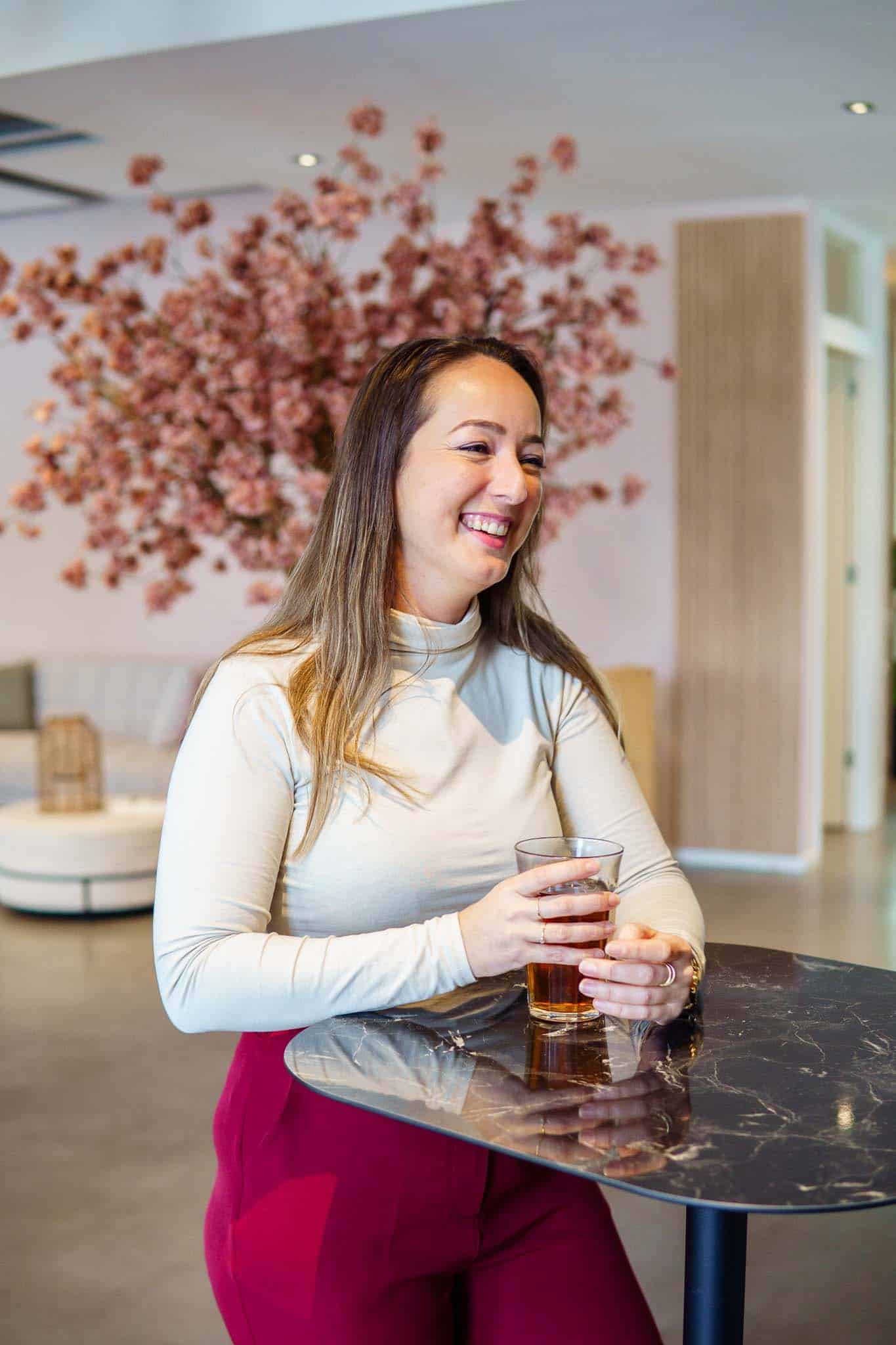 BURO210 Fullservice Marketingbureau Lachende vrouw geniet van een drankje aan een zwarte tafel in een stijlvolle lobby met bloemdecoratie op de achtergrond. Casual en ontspannen sfeer.