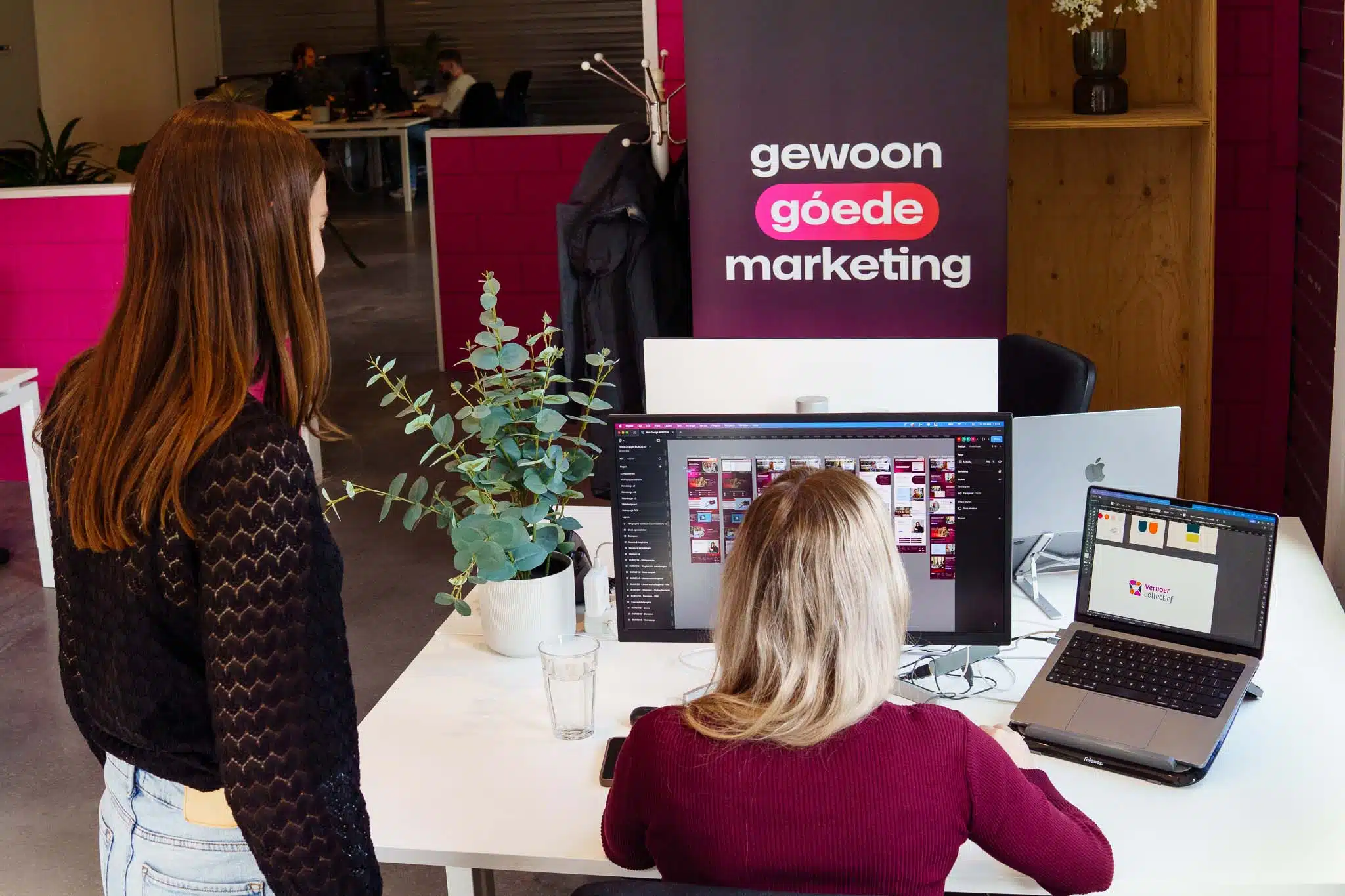 BURO210 Fullservice Marketingbureau Twee vrouwen werken samen op een kantoor van 'Gewoon Goede Marketing', met laptops en schermen vol grafisch ontwerp, omringd door moderne kantoordecoratie.