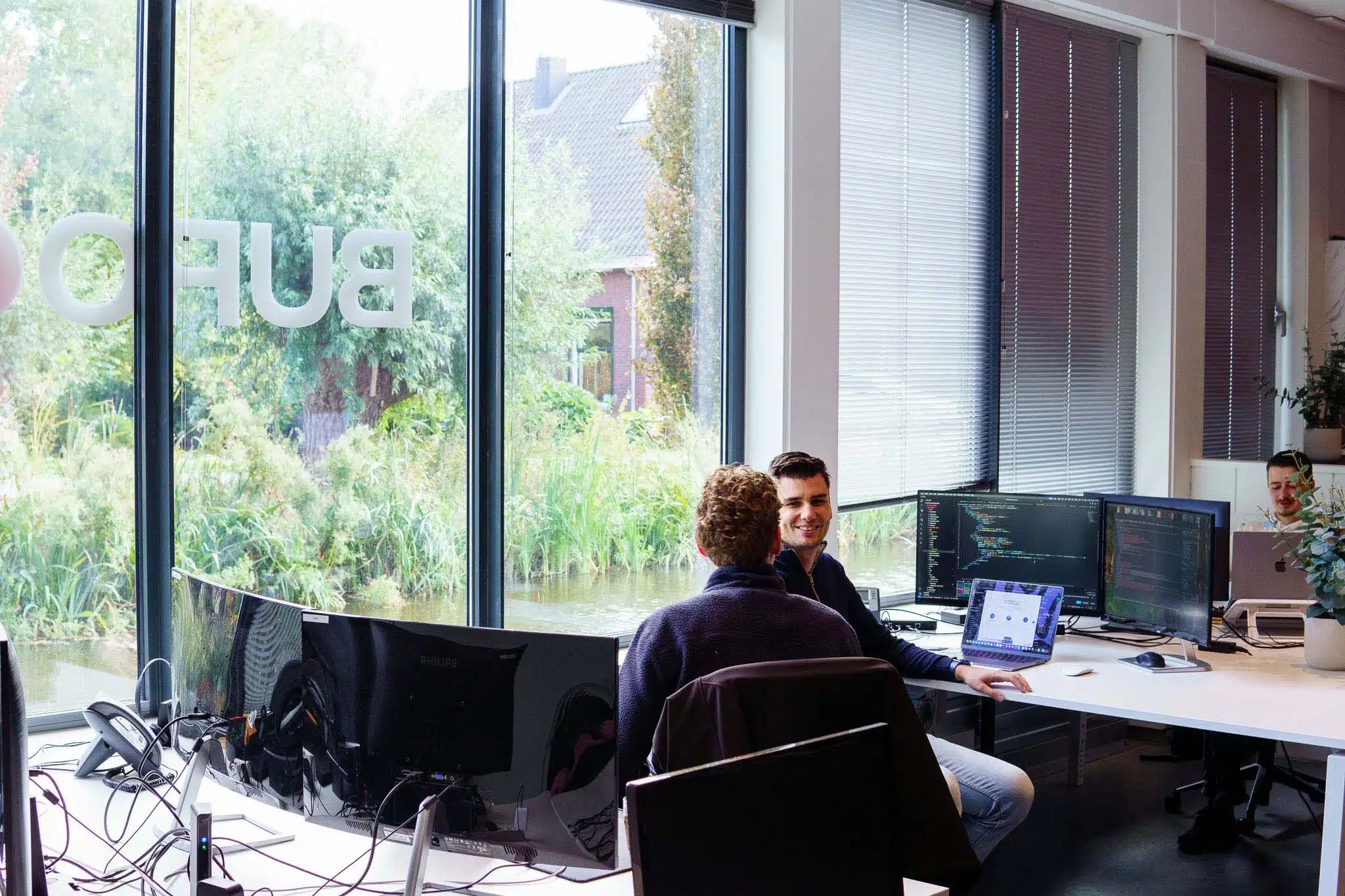 BURO210 Fullservice Marketingbureau Twee programmeurs werken samen in een modern kantoor met uitzicht op een rivier, computers met code, softwareontwikkeling en teamwerk.