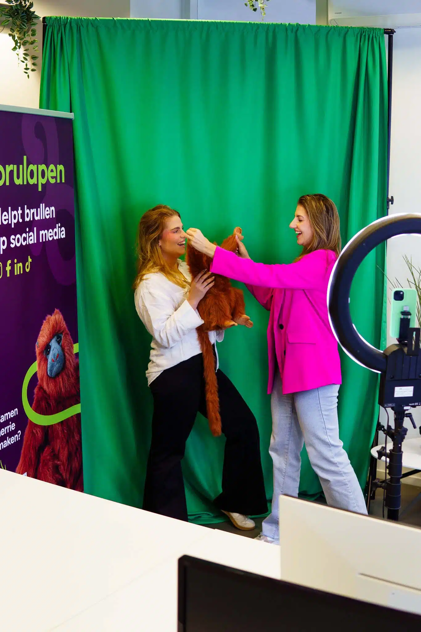 BURO210 Fullservice Marketingbureau Twee vrolijke vrouwen poseren met een aap voor een groene achtergrond, waarschijnlijk voor sociale media content creatie. Lachende gezichten en creativiteit in de spotlight.