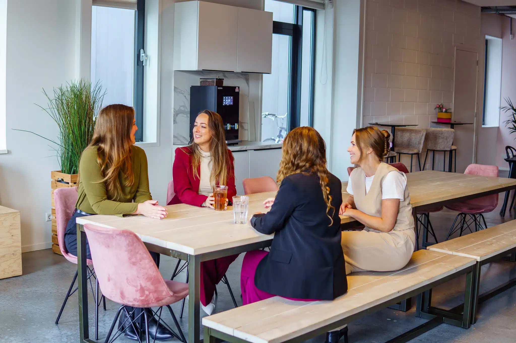 BURO210 Fullservice Marketingbureau Vier professionele vrouwen in een kantoorruimte zitten aan een tafel tijdens een informele bijeenkomst, genietend van elkaars gezelschap in een ontspannen sfeer.