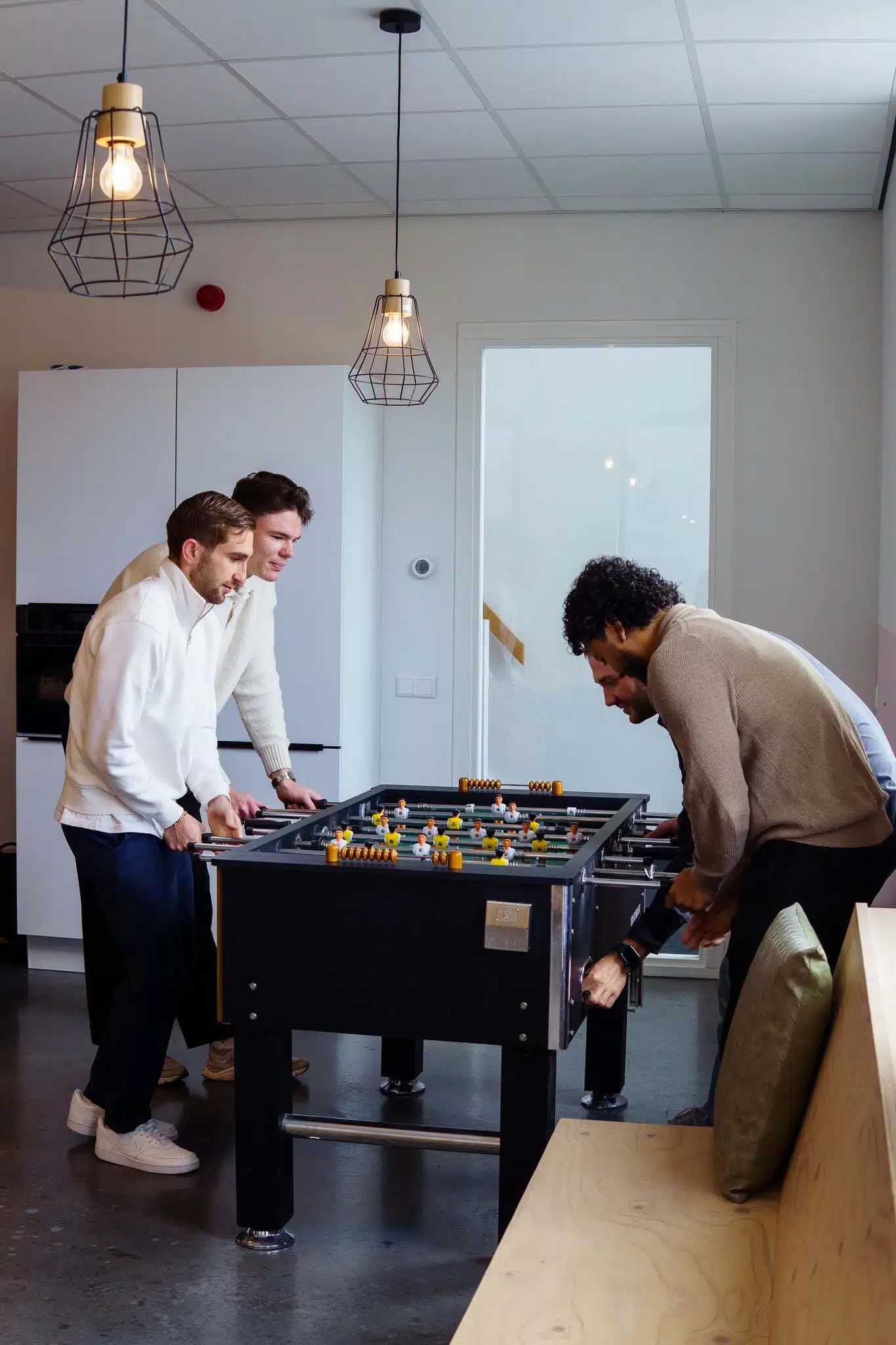 BURO210 Fullservice Marketingbureau Drie mannen spelen tafelvoetbal in een lichte, moderne kantooromgeving. De focus ligt op plezier en teambuilding tijdens een pauze.