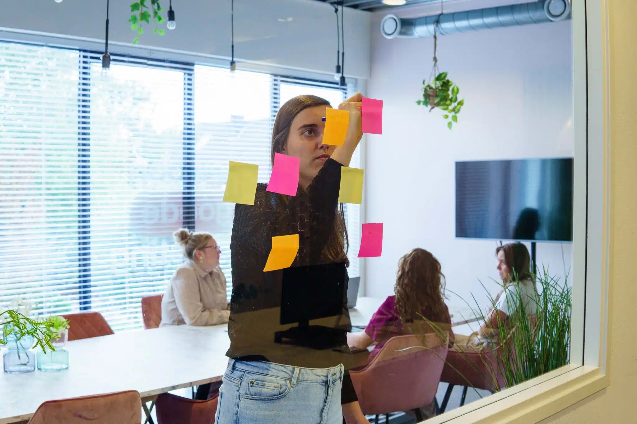 BURO210 Fullservice Marketingbureau Vrouw werkt samen met collega's aan een brainstormsessie, plakt kleurrijke post-its op een glazen wand in een moderne kantoorruimte voor planning en strategie.