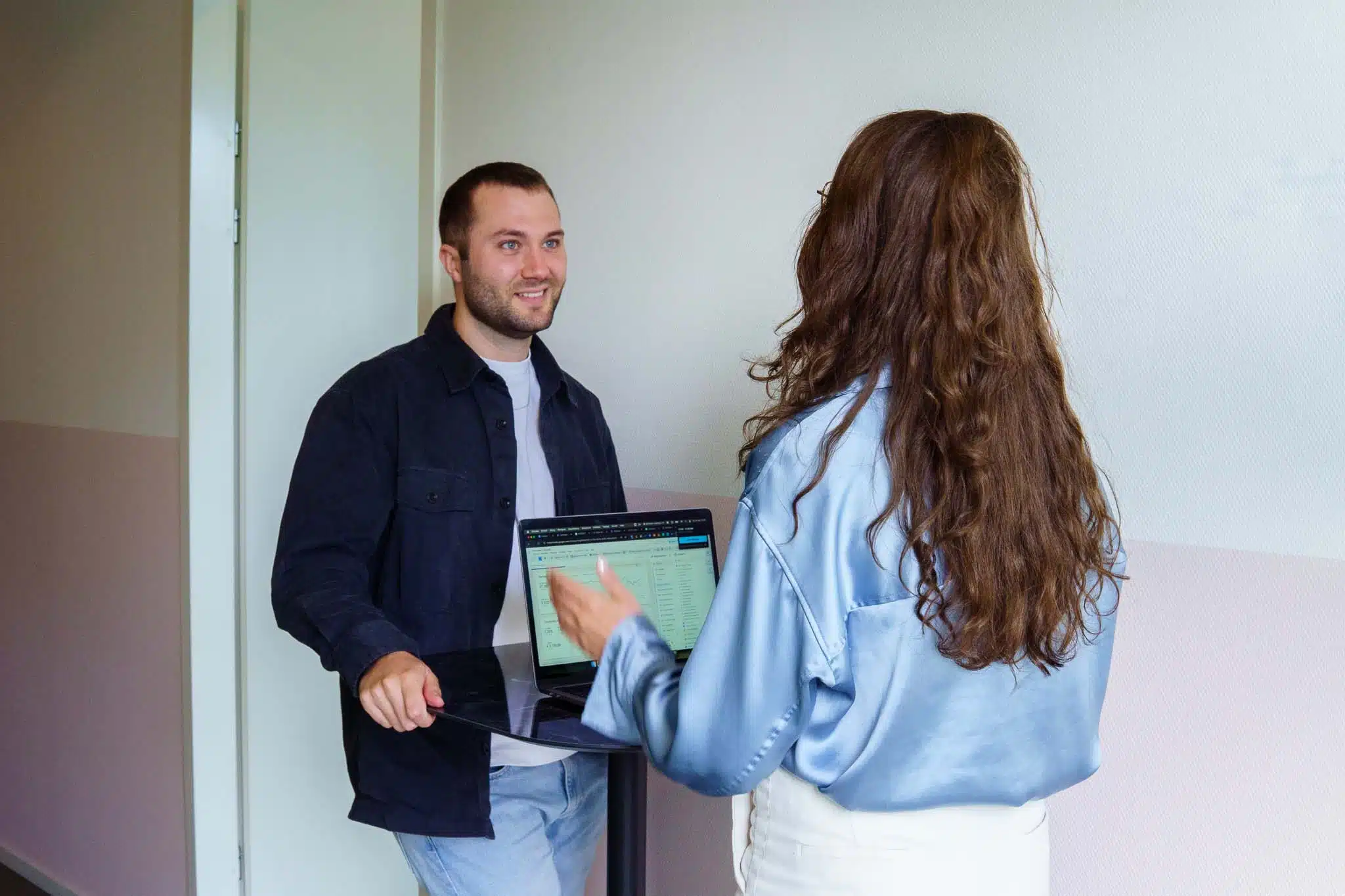 BURO210 Fullservice Marketingbureau Twee collega's bespreken data op een laptop in een moderne kantooromgeving, waarbij de man lacht en de vrouw uitleg geeft over grafieken en tabellen op het scherm.