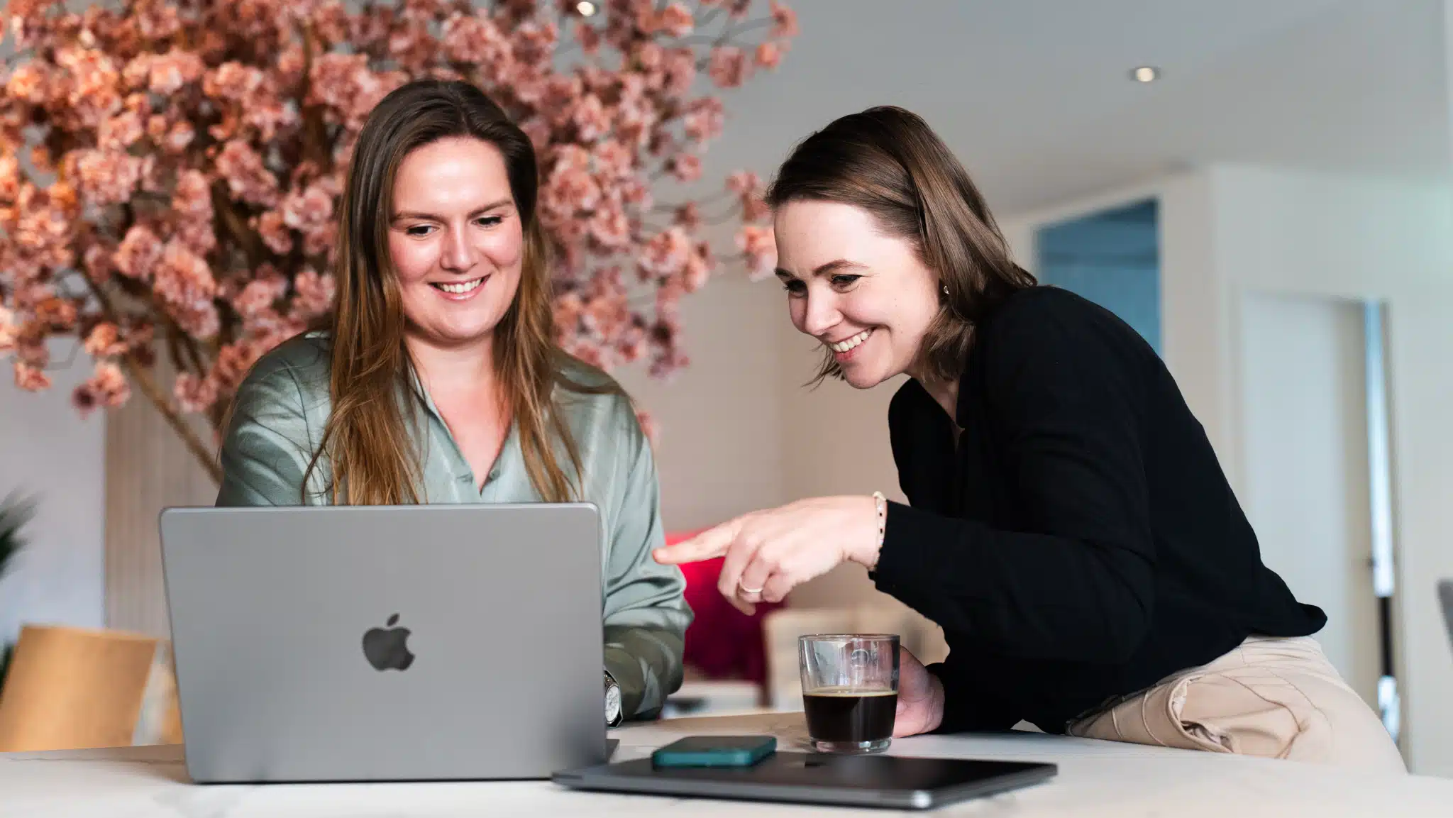 BURO210 Fullservice Marketingbureau Twee vrouwen werken samen aan een laptop, bespreken de visuele identiteit van een merk. Ze lachen en wijzen naar het scherm, wat een creatieve samenwerking suggereert.