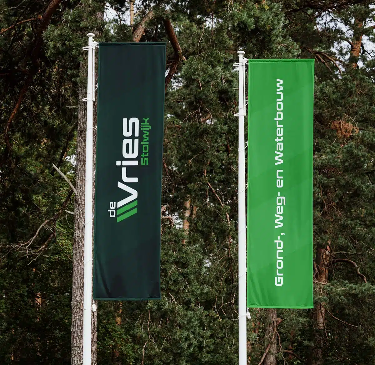 BURO210 Fullservice Marketingbureau Twee vlaggen van De Vries Stolwijk, een donkergroene en een lichtgroene, wapperend voor bomen. Perfect voor grond-, weg- en waterbouw projecten.